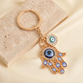 Gold tone openwork Hamsa keychain on a cream background, with a large blue palm eye, smaller finger eyes and a round side charm.