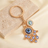 Gold tone openwork Hamsa keychain on a cream background, with a large blue palm eye, smaller finger eyes and a round side charm.