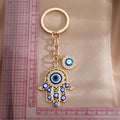 Gold tone openwork Hamsa keychain with blue eye details, laid vertically beside a ruler.