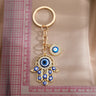 Gold tone openwork Hamsa keychain with blue eye details, laid vertically beside a ruler.