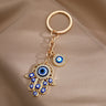 Gold tone openwork Hamsa keychain on taupe fabric, showing blue eyes across the hand and a round eye charm beside the chain.