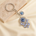 Silver tone openwork Hamsa keychain on a cream background, showing blue eye motifs, clear accents and the short link chain.