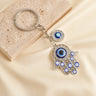 Silver tone openwork Hamsa keychain on a cream background, showing blue eye motifs, clear accents and the short link chain.