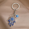 Silver tone openwork Hamsa keychain on taupe fabric, showing blue eyes across the hand and a round eye charm beside the chain.
