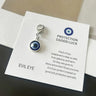 Close view of a blue round evil eye keychain charm with a silver tone rim, connecting links and lobster clasp on a printed card.