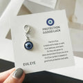 A hand holds a printed card displaying the Blue evil eye keychain charm with concentric colored rings and a silver tone lobster clasp.