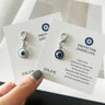 A hand holds two printed cards displaying white and blue evil eye keychain charms with silver tone frames and lobster clasps.