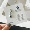 A hand holds a printed card displaying the White evil eye keychain charm with a light blue ring, dark center and silver tone lobster clasp.