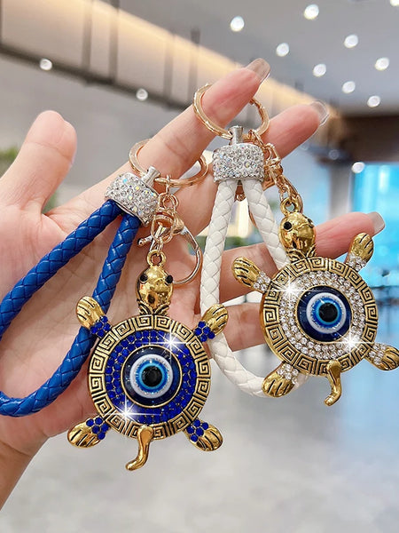 Blue and white turtle evil eye keychains held side by side, with braided loops and gold tone patterned shells.