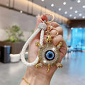 White turtle evil eye keychain held upright, showing clear accents around the blue eye, a white braided loop and gold tone hardware.
