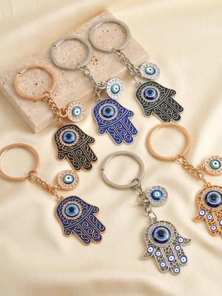 Six Hamsa evil eye keychain designs, showing blue and black scroll hands and openwork hands with gold tone or silver tone details.