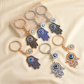 Six Hamsa evil eye keychain designs, showing blue and black scroll hands and openwork hands with gold tone or silver tone details.
