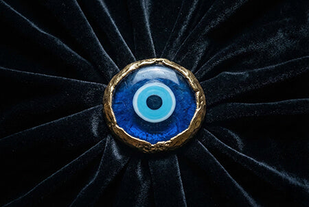 A close-up photograph of a large, mysterious blue glass evil eye amulet with a rugged gold rim, resting centrally on dark folds of velvet fabric.