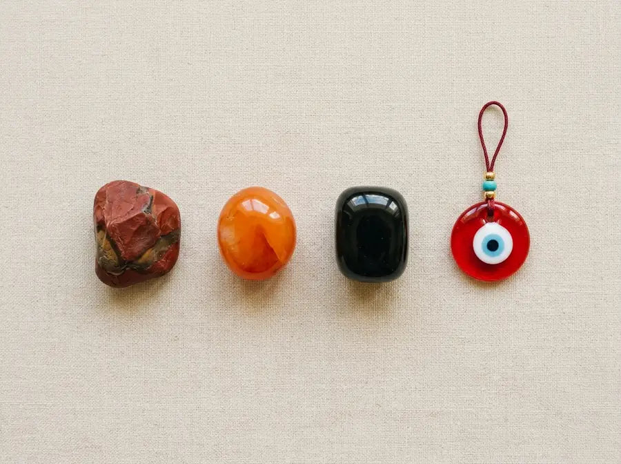 Red jasper, carnelian, and black obsidian stones arranged in a row beside a red evil eye charm on a warm surface
