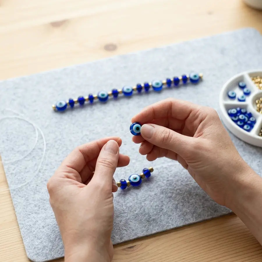 Hands threading one evil eye bead onto clear cord while the remaining planned bead row stays aligned behind the work area.