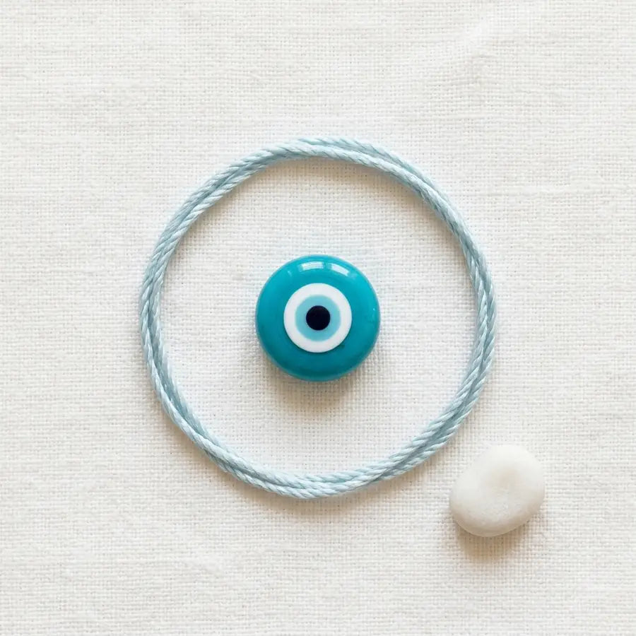 Single turquoise evil eye bead inside a pale-blue cord circle on white linen with a small white pebble nearby.