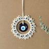 Blue evil eye disc suspended inside an off-white fringed circle with small eye beads, hanging against a tan background.
