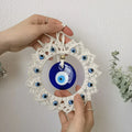 Two hands holding the loop and lower frame of the blue and off-white evil eye wall hanging at a slight angle.