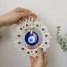 Two hands holding the loop and lower frame of the blue and off-white evil eye wall hanging at a slight angle.