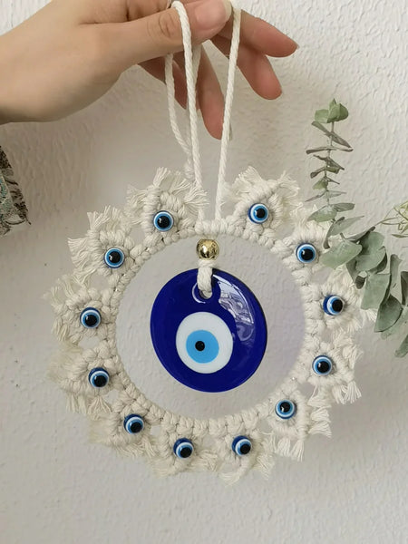 Hand holding the off-white cord loop of a circular blue evil eye wall hanging against a white wall beside greenery.