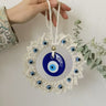 Hand holding the off-white cord loop of a circular blue evil eye wall hanging against a white wall beside greenery.