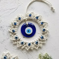 Circular blue evil eye wall hanging lying on a pale textured surface, with its off-white hanging loop arranged above the fringed frame.