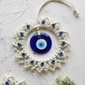 Circular blue evil eye wall hanging lying on a pale textured surface, with its off-white hanging loop arranged above the fringed frame.