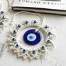 Angled view of the blue evil eye wall hanging on a white textured surface beside a book, showing raised border beads and off-white fringe.