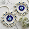 Closer angled photograph of two blue evil eye wall hangings with off-white beaded fringe beside white flowers.