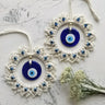 Two blue and off-white circular evil eye wall hangings arranged diagonally on a pale textured surface beside white flowers.